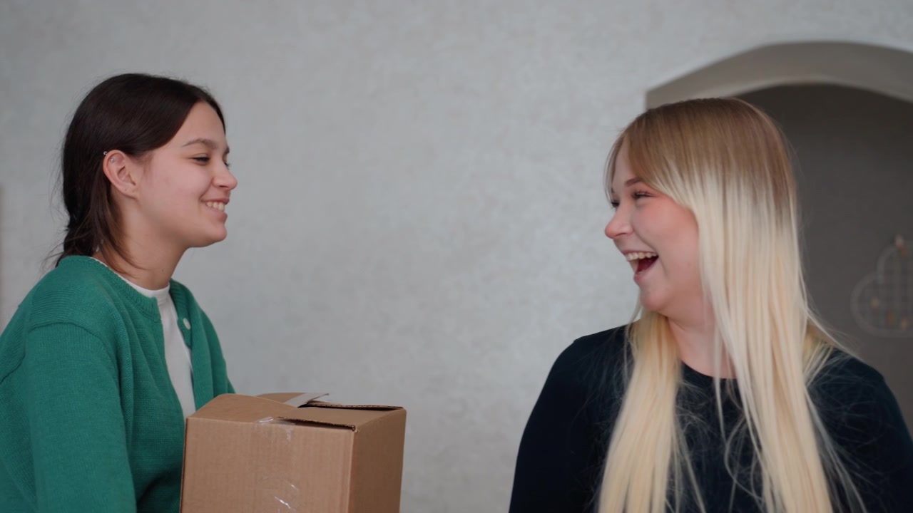 Donne che ridono con una pianta e una scatola all'interno di un appartamento accogliente, la bionda tiene una pianta succulenta mentre la mora porta del cartone, sorrisi giocosi e conversazione calorosa, luce solare su una parete d'accento verde acqua, supporto condiviso.
