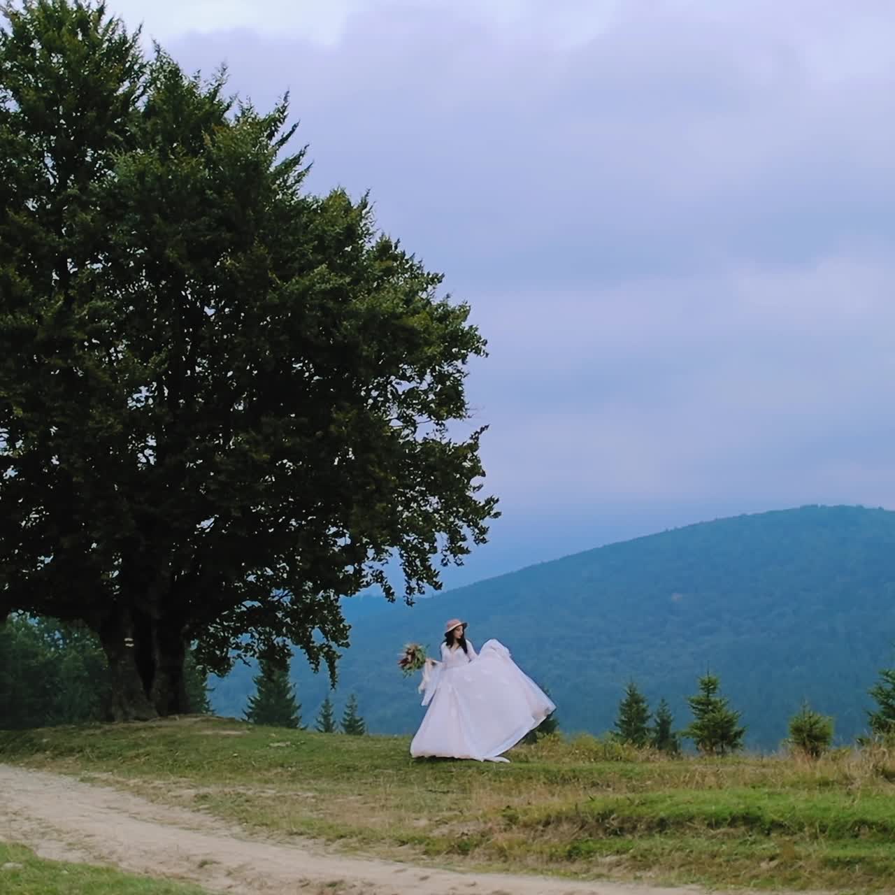 Woman in wedding dress on nature background. Young bride in white dress and hat standing near the green tree in the mountains.