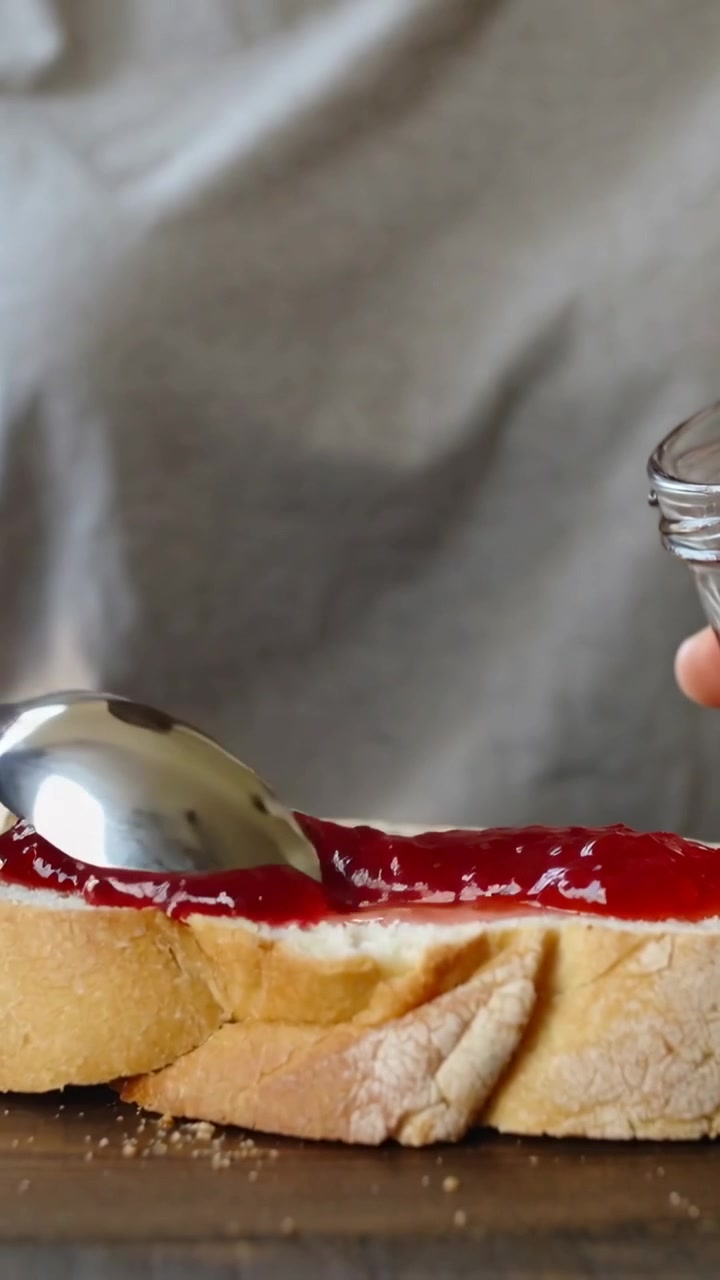 Vertical video: Dipping spoon, hand spreading jam across bread for snack with jar, in grey shirt