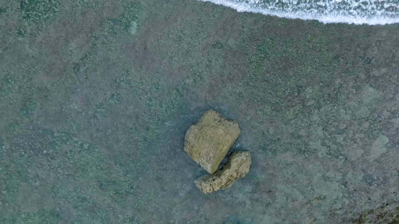 Top Down Slow Motion Drone View of Waves crashing over shallow coral reef and big rock in Uluwatu Bali Indonesia