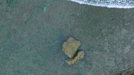 Top-Down-Slow-Motion-Drone-View-of-Waves-crashing-over-shallow-coral-reef-and-big-rock-in-Uluwatu-Bali-Indonesia