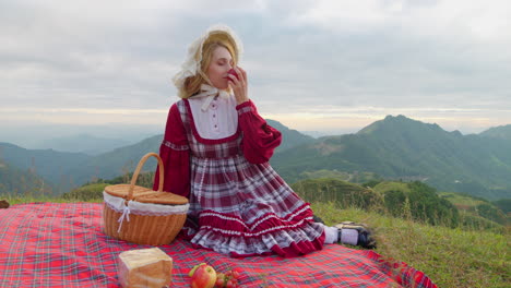 young blonde female model in traditional old fashioned renaissance dress clothing sitting alone having lunch and eating a red apple in mountains landscape historical reproduction