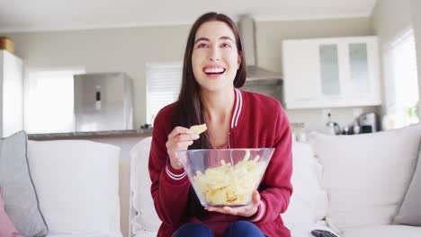 happy caucasian woman sitting on couch, eating snacks and watching tv in living room