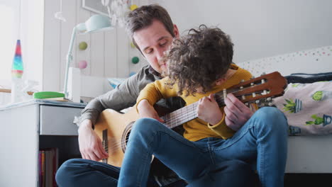 single father at home with son teaching him to play acoustic guitar in bedroom