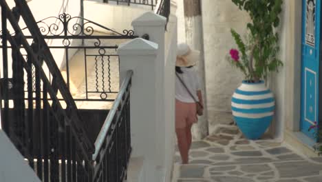 Chica-En-Traje-De-Senderismo-Veraniego-Caminando-Por-El-Típico-Callejón-Griego-Estrecho-Con-Puertas-Azules-Y-Plantas