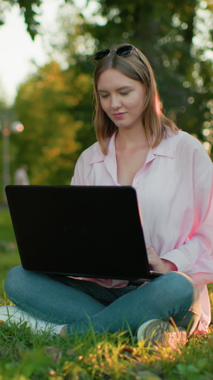 Freelancer in pink top with glasses resting on her head smiles warmly and continues typing on her laptop while sitting outdoors, trees and people walking appear in background with natural lighting