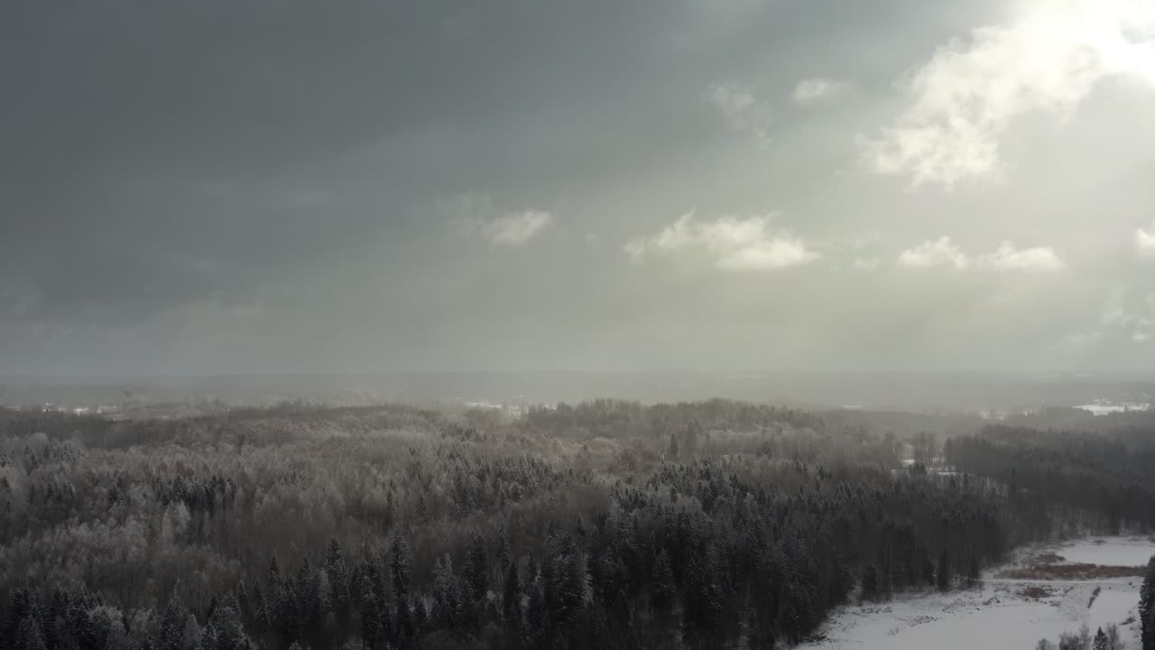 Aerial drone view of a dramatic winter sky with sun rays piercing through clouds over a snow-covered forest in countryside.