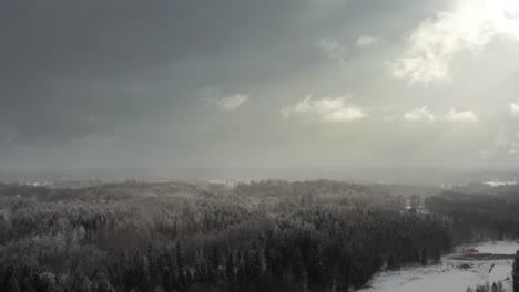 aerial drone view of a dramatic winter sky with sun rays piercing through clouds over a snow-covered forest in countryside.