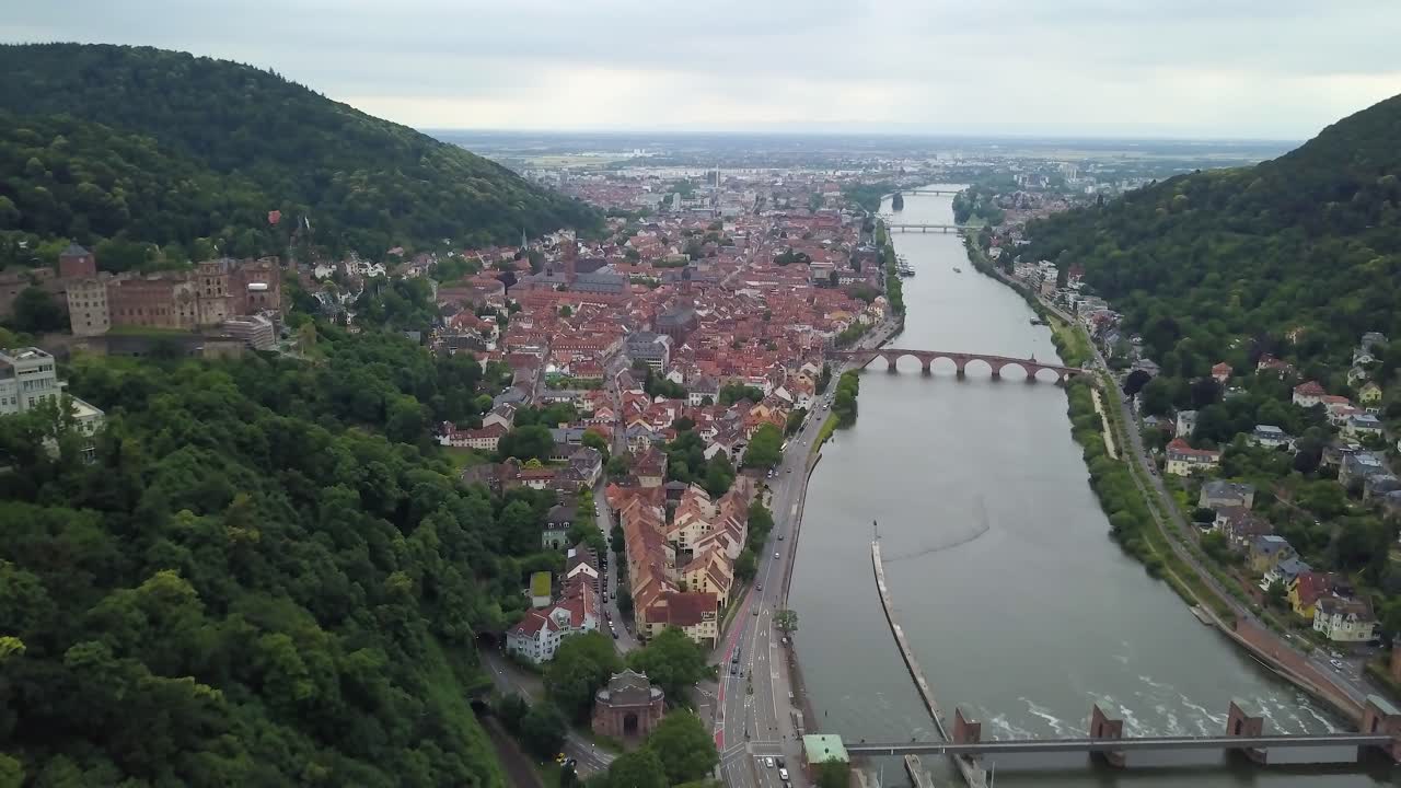 Dolly out aereo del villaggio e delle rovine del Castello di Heidelberg, vicino al fiume Neckar e ai suoi ponti, tra colline coperte da fitta foresta verde, Germania