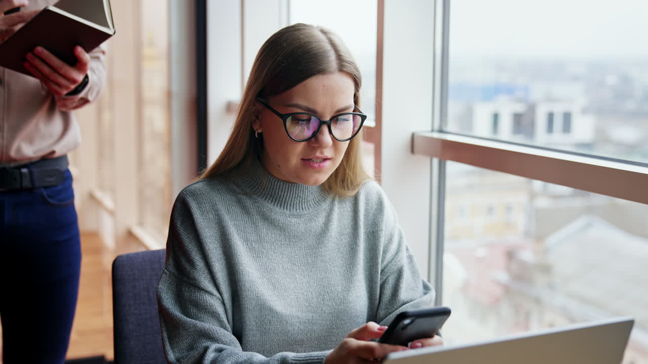 Pretty woman in glasses takes a phone in hands. Lady is looking for a contact talking to a man standing behind.