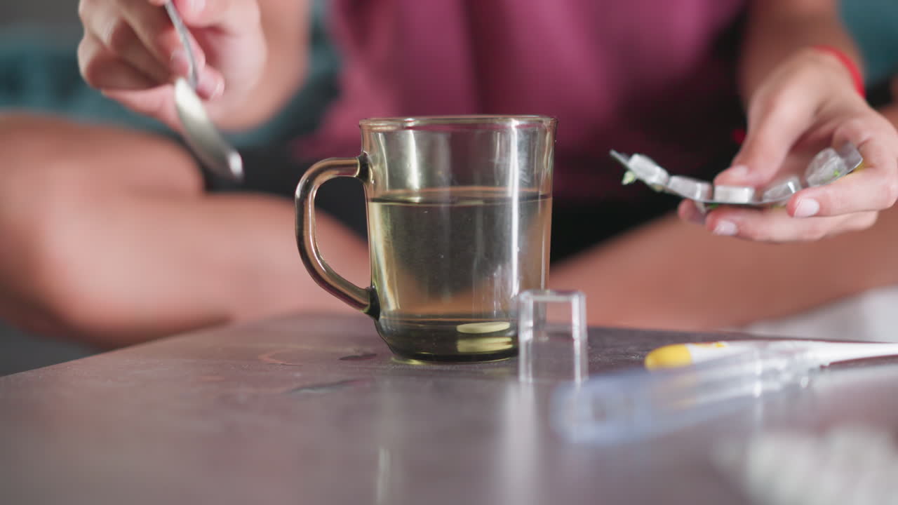 Close up of young girl preparing medicine by pouring drug into warm water in glass cup with spoon nearby while seated on couch caring for sick brother during home treatment