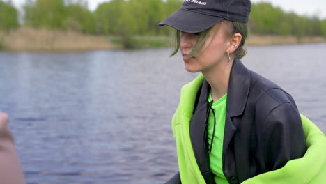 jeune femme avec une casquette noire et une veste en cuir tenant un appareil photo à côté de bouteilles de bière au bord de la rivière