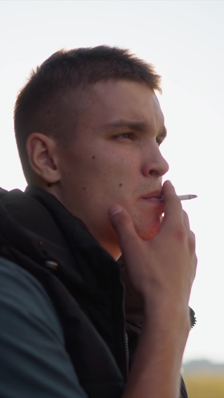 Primer plano de un hombre sosteniendo y fumando un objeto con iluminación suave. El sujeto viste ropa oscura, los detalles visibles de la textura de la piel incluyen pecas en el cuello. El fondo muestra un cielo brillante y elementos distantes