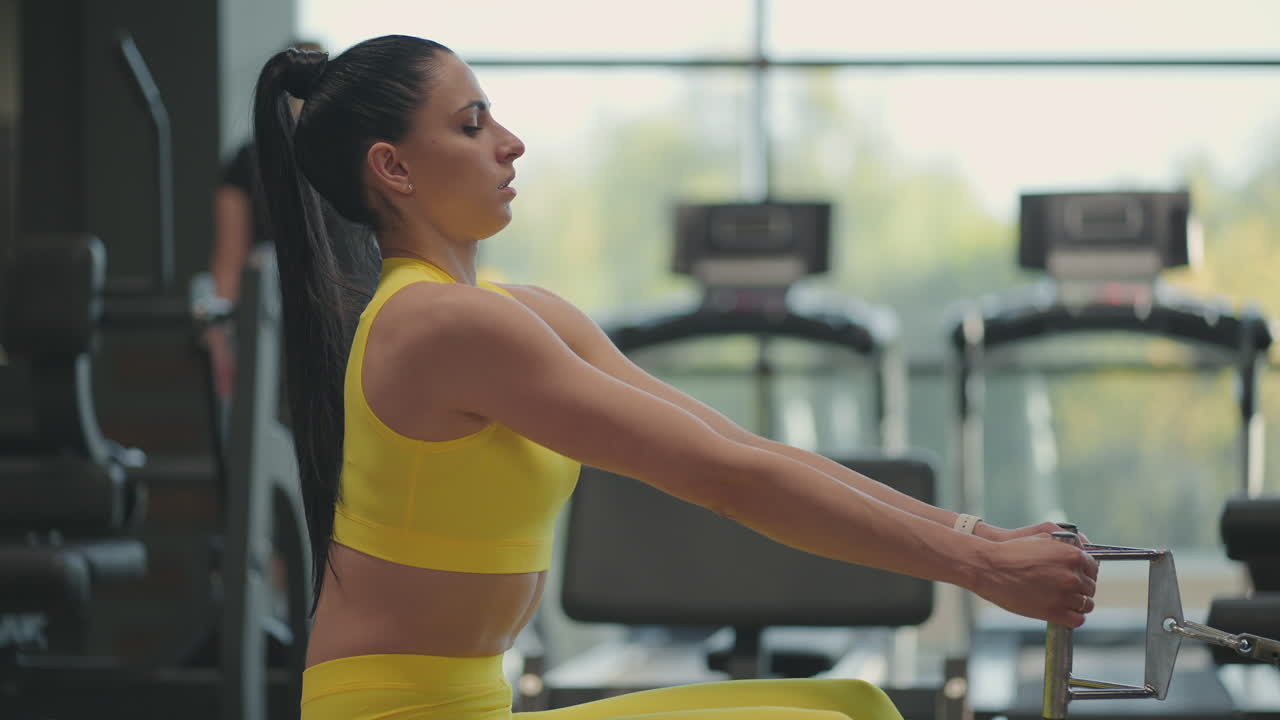 una mujer hispana hace ejercicios en la máquina de remo. una chica se sienta detrás de un simulador se llama una máquina de remo. sentada tira del peso de las grandes ventanas y cintas de correr del gimnasio