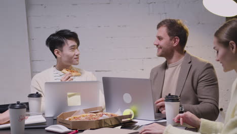 two multiethnic colleagues eating pizza and talking to each other during a team meeting in the boardroom