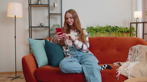 Cheerful-young-woman-sitting-on-sofa,-using-mobile-phone-share-messages-on-social-media-application