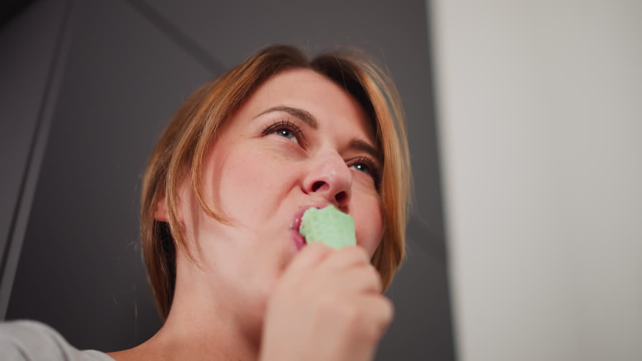 Close up upward view of elegant girl biting into ice cream cone, savoring sweet treat with delight, standing indoors against minimalist background