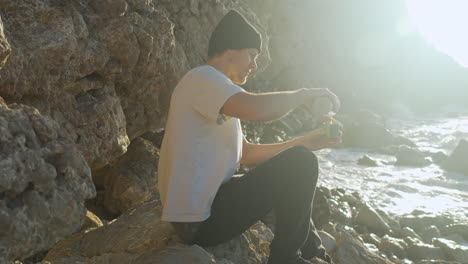 man unscrews thermos, pours coffee in cup, drinks it, as morning sun shines on him while sea waves crush into rocks near by