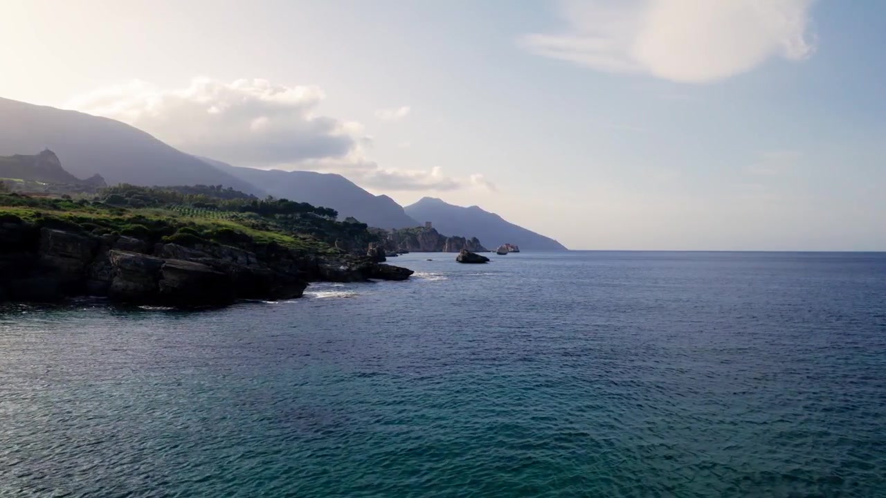 Una luce dorata e soffusa e ombre di nuvole che si muovono si spostano su un mare calmo accanto a scogliere rocciose sulla costa settentrionale della Sicilia