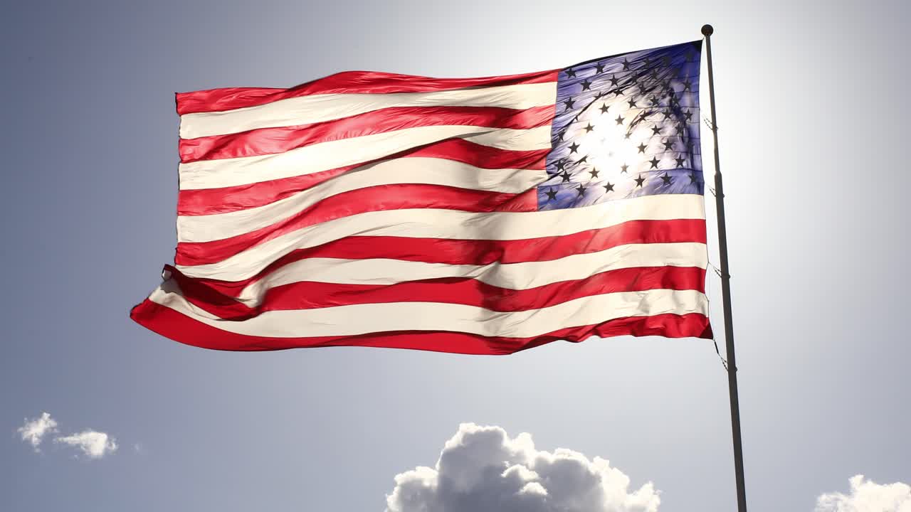 US Flag Waves in the Wind High Above the Ground with Sunlight Behind on a Blue Sky Day with Clouds. American Flag with Stars and Stripes for Presidential Elections and Patriotism for United States.