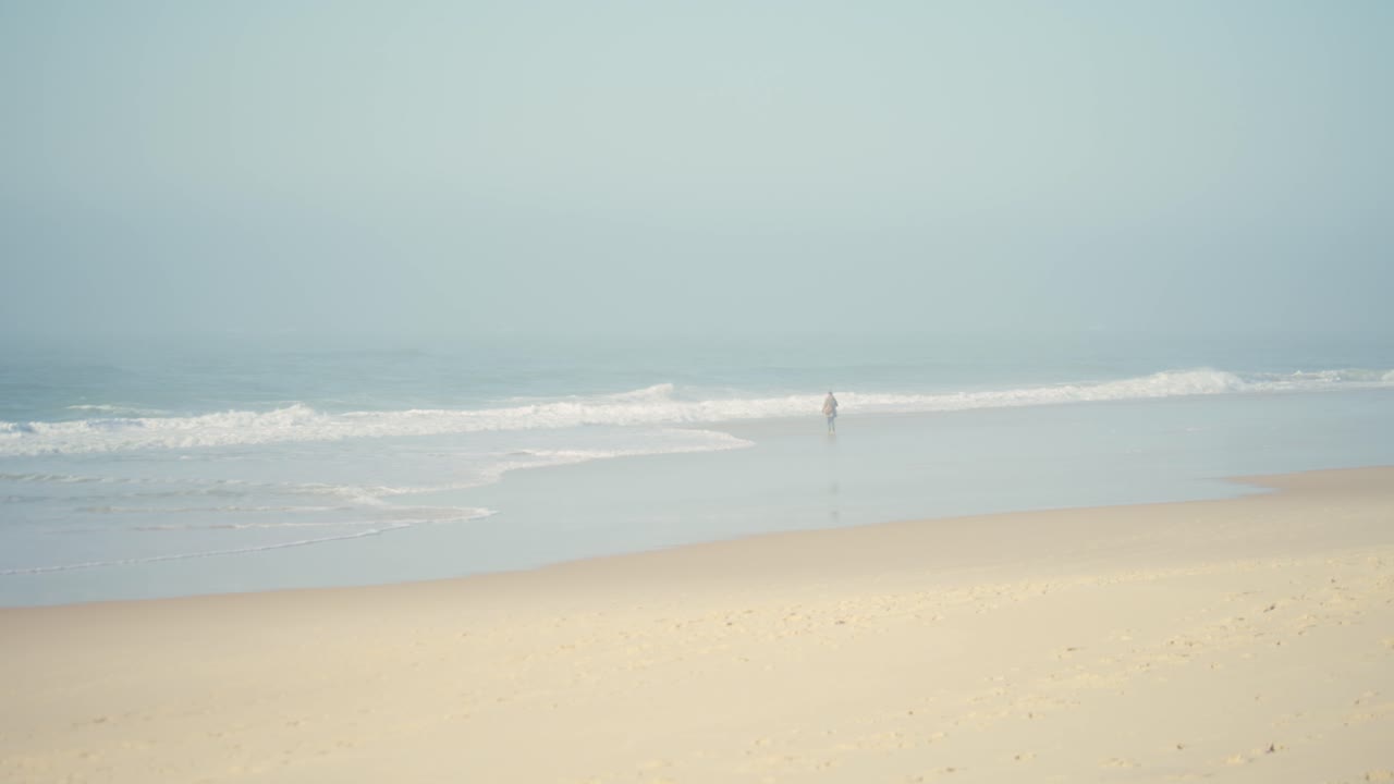 Unrecognizable person walking alone on misty beach at dusk. Ocean waves rolling in. Static long shot