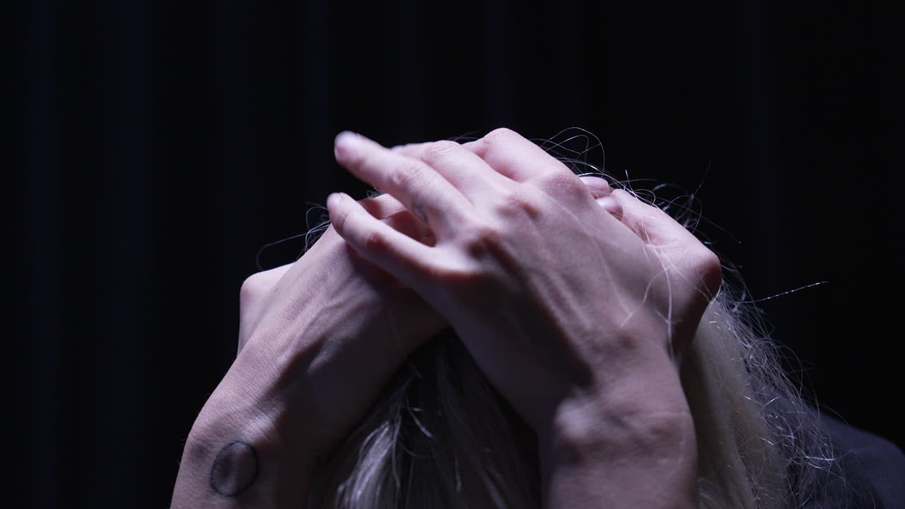 손으로 머리를 감싸고 있는 사람, 하나의 머리 위 전등이 비추는 어두운 방에서 손가락이 초조하게 안절부절 못하고 있습니다.