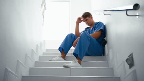 stressed and tired male doctor or nurse wearing scrubs sitting on stairs in hospital
