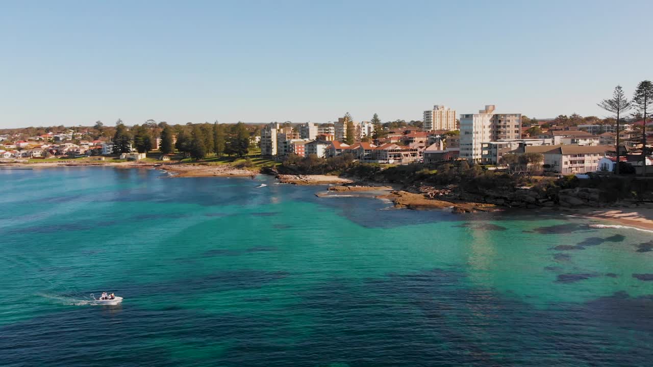 생생한 푸른 물의 드론 뷰, 그것은 상승으로 해안으로 돌아보고, 도시의 광범위한 을 하기 전에 배경에서 지나가는 모터 보트와 함께