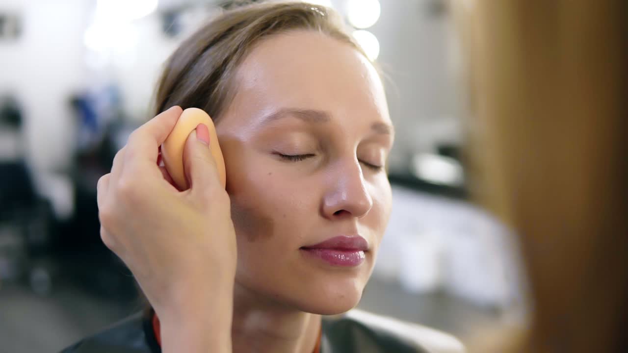 The make-up artist puts contours on cheekbones of the girl. Professional makeup. Young, blonde model