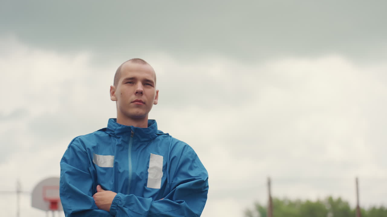 Hombre blanco con impermeable azul en una cancha exterior, retrato estoico de un entrenador bajo nubes de tormenta, con los brazos cruzados, canasta de baloncesto y valla de tela metálica al fondo, cámara lenta cinematográfica, gradación de color sombría, resiliente