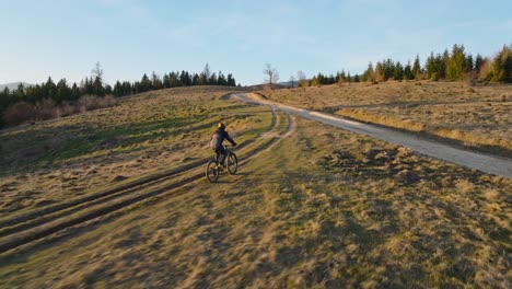 cyclist riding along a dirt trail in an open field surrounded by trees at sunset