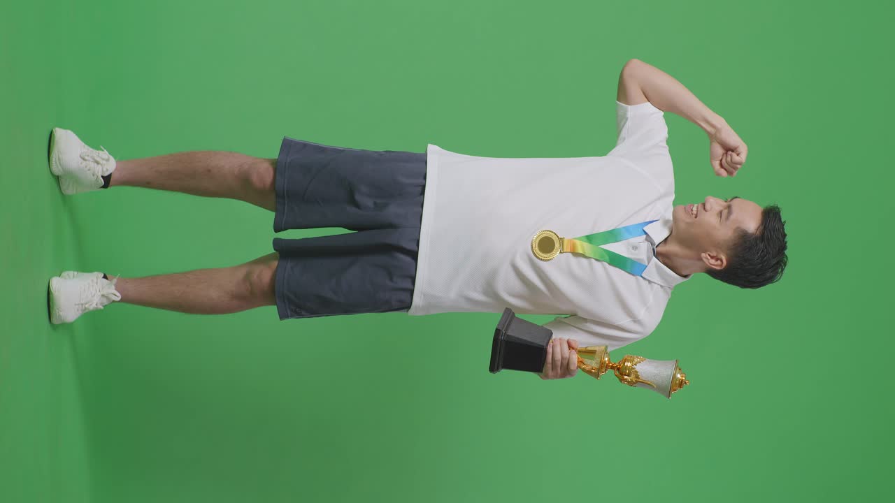 Full Body Of Asian Man With A Gold Medal And Trophy Flexing His Bicep And Smiling To Camera On Green Screen Background In The Studio