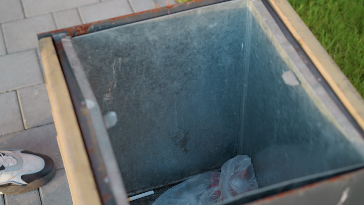 close up of urban trash bin interior showing metal liner, plastic bag and debris at bottom, with sneakered foot of person standing on paved sidewalk beside grassy verge