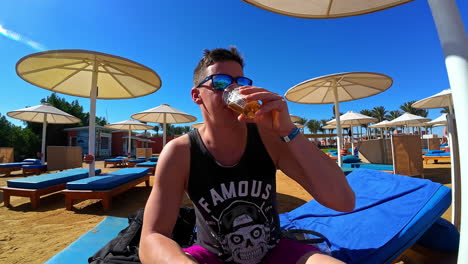 Man-enjoying-a-cold-drink-at-a-sunny-resort-with-blue-loungers-and-parasols,-Egypt