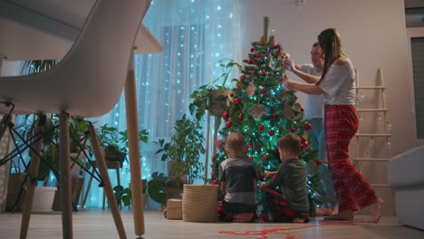 Familia-Feliz-Preparándose-Para-El-Concepto-De-Celebración-De-Las-Vacaciones-De-Invierno-De-Año-Nuevo.-Una-Pareja-Joven-De-30-Años,-Sus-Adorables-Dos-Hijos-Preescolares-Decorando-El-árbol-De-Navidad-Crean-Un-Ambiente-Festivo-En-Una-Casa-Moderna-Y-Acogedora.-Imágenes-4k-De-Alta-Calidad