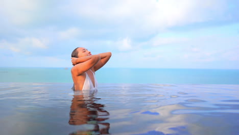 sensual joven asiática toca su cabello mojado estando dentro del agua de la piscina infinita en la azotea de un hotel de lujo con paisaje marino en el fondo