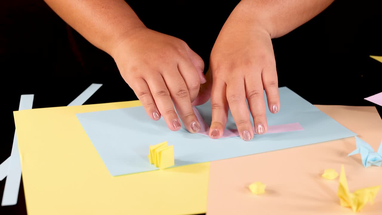 Person folds pastel origami paper on a bright table, overhead view, even studio lighting