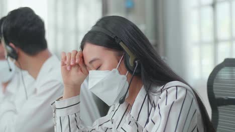 アジア人女性コールセンターエージェントがヘッドセットとマスクをかぶって頭痛を感じています彼女の2人の同僚がオフィスで働いている間話しているからです
