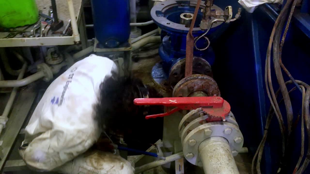 Man with long hair in white jumpsuit sawing a rusty valve while reparing a boat