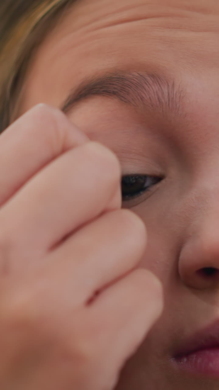 Primer plano de una adolescente poniéndose pestañas postizas, dedos firmes con uñas brillantes presionando la tira de pestañas sobre el párpado superior, luz natural suave, expresión concentrada, rutina de belleza íntima