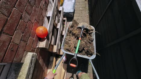 pov of man wheeling a wheelbarrow full of soil and a spade down the side of a house on a sunny day