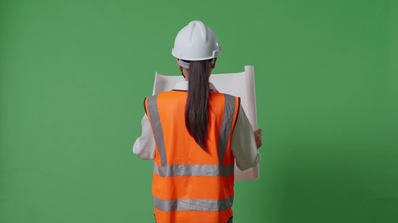 안전 헬을 착용한 아시아 여성 엔지니어가 손에 청사진을 들고 녹색 화면 배경 스튜디오에서 는 동안 주변을 둘러보고 있습니다.