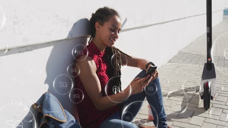 young woman sitting against wall using smartphone with 5g network animation overlay
