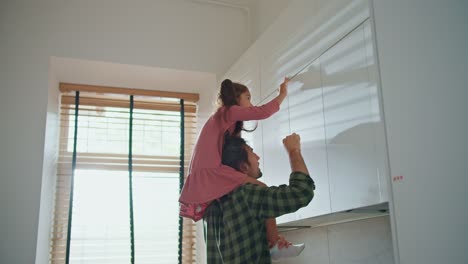 A-little-brunette-girl-in-a-pink-dress-helps-her-brunette-dad-in-a-plaid-shirt-get-an-item-from-the-top-shelves-in-the-kitchen.-A-little-girl-sits-on-her-father's-shoulders-and-takes-an-object-from-a-high-shelf-in-the-kitchen