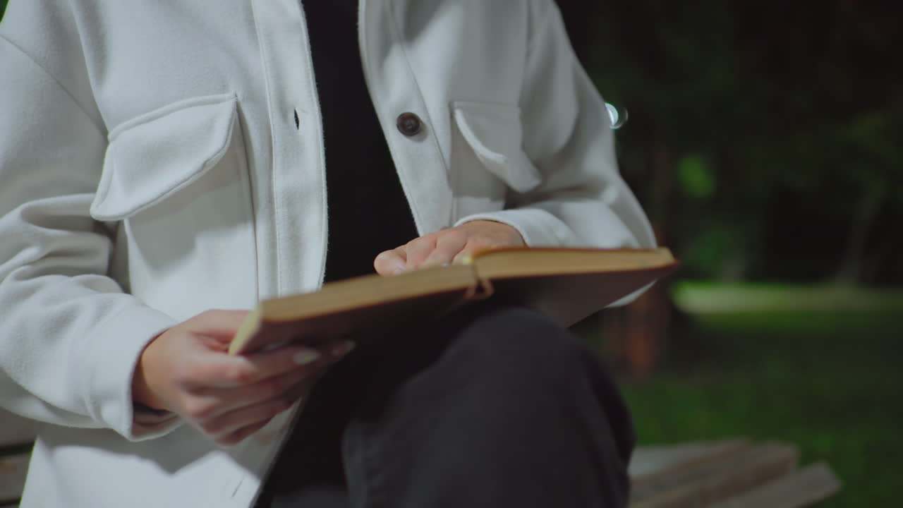 Partial view of lady sitting outdoors at night on wooden bench opening to another page of book under soft streetlight with blurry trees and dim natural background