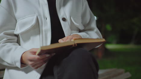 partial view of lady sitting outdoors at night on wooden bench opening to another page of book under soft streetlight with blurry trees and dim natural background