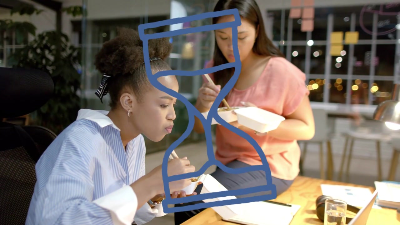 Two female coworkers in office working late, eating takeout, checking laptop while hourglass hiding