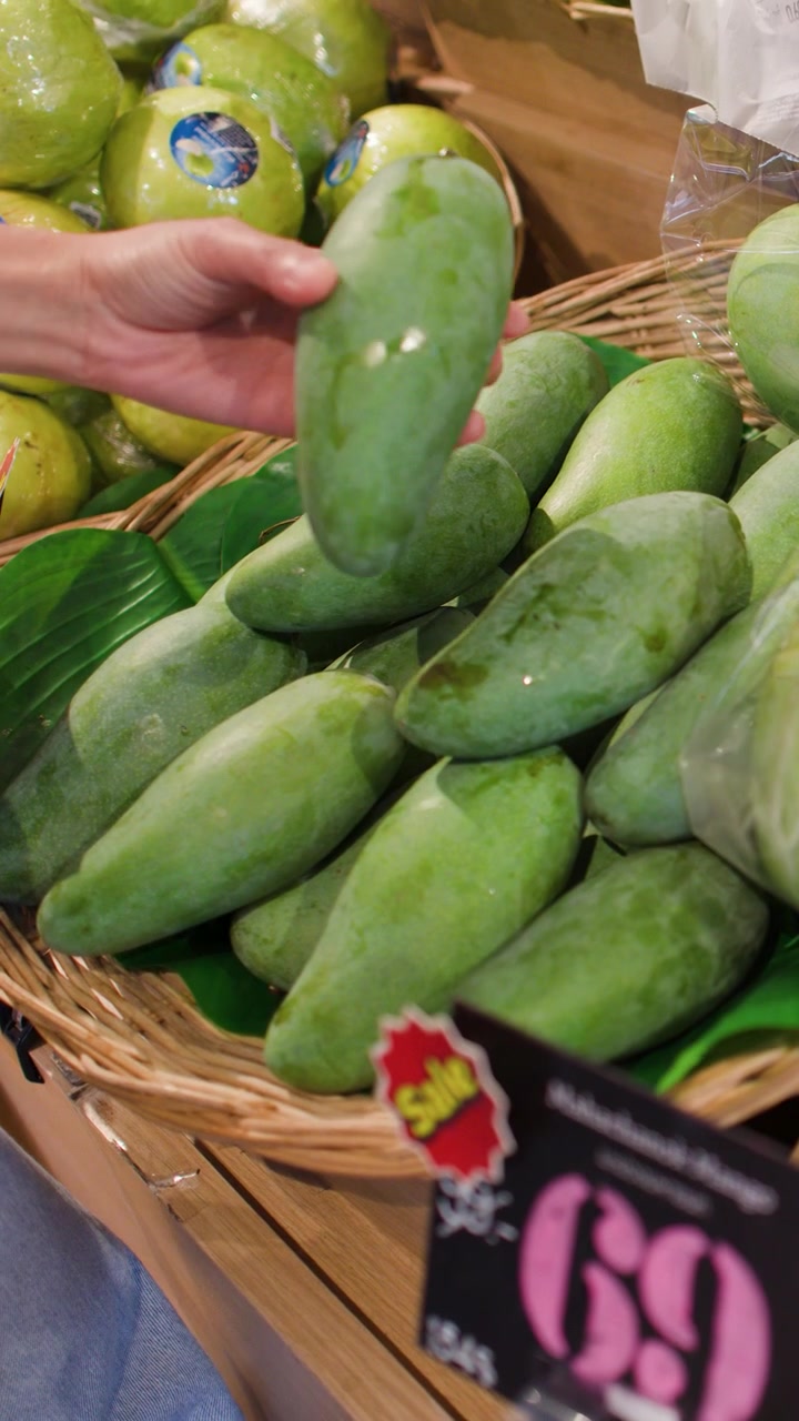 Una mano ispeziona e seleziona attentamente manghi verdi freschi da un espositore di un negozio di alimentari.