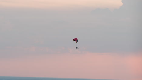 distant view of colorful paraglider soaring above serene horizon at sunset, silhouetted pilot suspended beneath curved canopy against soft pastel sky gradient conveying tranquil aerial adventure