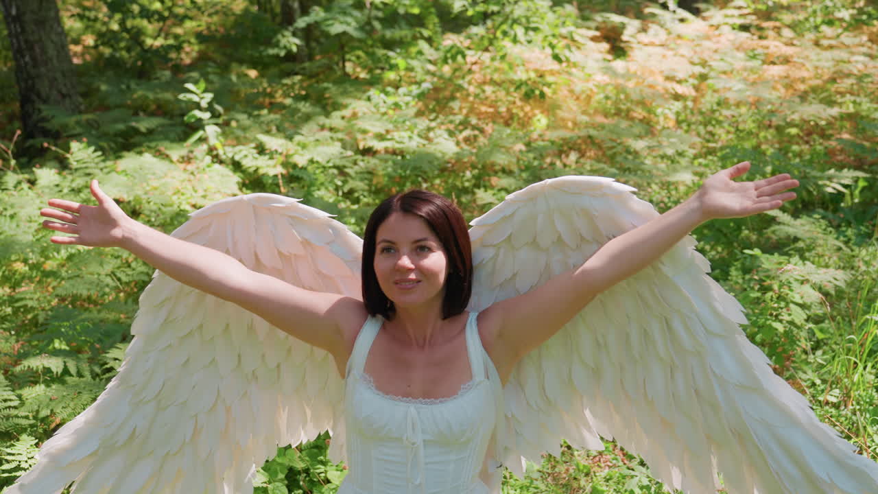 Young lady wearing costume wings lifts hands and embraces warmth of nature in lush green forest as sunlight glows gently around her, creating peaceful ethereal scene filled with love
